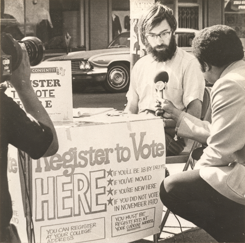 Picture of David Mundstock at streetside voter registration table being interviewed by TV media, 1971.